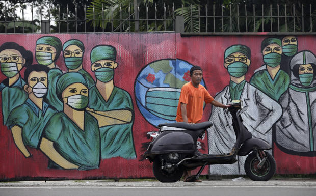 Dalam foto tertanggal 15 April 2020 ini, seorang lelaki dengan kendaraan bermotornya berdiri di depan sebuah mural yang dibuat untuk menghormati para tenaga kesehatan yang telah banyak berjatuhan dalam penanganan pandemi COVID-19. (AP)