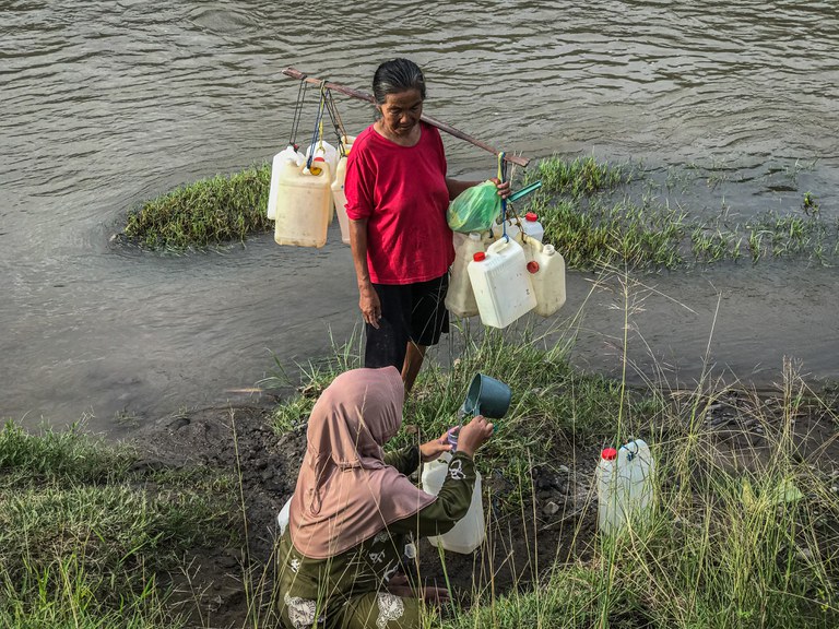 Nenek Samriati berdiri sambil memikul jeriken kosong menunggu keponakannya Indar Sari selesai mengisi air bersih ke dalam jeriken di pinggir sungai Desa Renggeang, Kecamatan Limboro, Kabupaten Polewali Mandar, Provinsi Sulawesi Barat, 11 Mei 2022. Hujan deras sejak dua hari terakhir di desa itu membuat air sungai meluap sehingga menyulitkan warga mendapatkan titik untuk penggalian mata air. Ketika satu mata air ditemukan, warga harus antre untuk bisa menggunakannya mengambil air bersih. [Keisyah Aprilia/BenarNews]