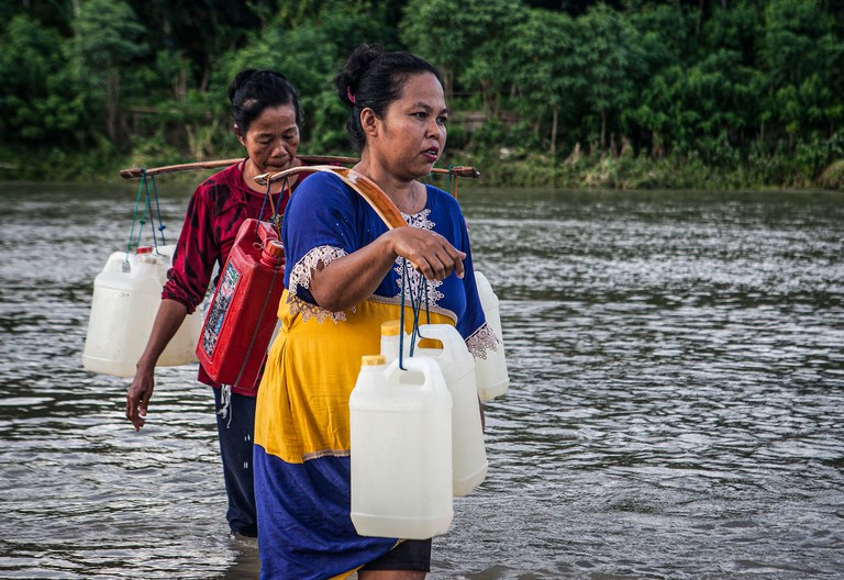 Dua warga berjalan menyeberangi sungai sambil memikul jeriken berisi air bersih di sungai Desa Renggeang, Kecamatan Limboro, Kabupaten Polewali Mandar, Provinsi Sulawesi Barat, 11 Mei 2022. Sebagian warga yang bermukim di seberang sungai terpaksa harus menyeberangi sungai untuk mendapatkan air bersih. Air sungai tidak dalam sehingga tidak membahayakan mereka. Mayoritas warga di desa ini berjalan kaki untuk mengambil air bersih di sungai Renggeang. Meski jaraknya jauh antara tiga sampai empat kilo meter dari rumah mereka, warga tidak menjadikan itu beban. Menurut mereka ketimbang harus mengeluarkan uang untuk membeli air bersih di pasar mending mereka mengambilnya di sungai secara gratis. [Keisyah Aprilia/BenarNews]