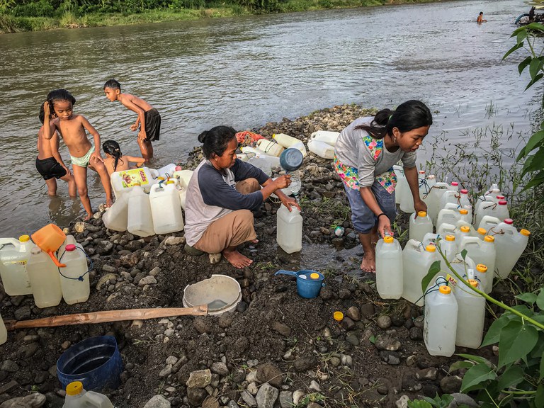 Ratna Jani (duduk) mengisi air bersih ke dalam jeriken sementara anaknya, Susan Jani, mengatur jeriken yang sudah terisi air bersih di pinggir sungai Desa Renggeang, Kecamatan Limboro, Kabupaten Polewali Mandar, Provinsi Sulawesi Barat, 11 Mei 2022. Ibu dan anak ini setiap sore mengambil air bersih di sungai untuk dijual kepada warga lainnya di desa seharga Rp1.000 per jeriken.  Tampak di latar belakang anak-anak bermain di sungai. [Keisyah Aprilia/BenarNews]