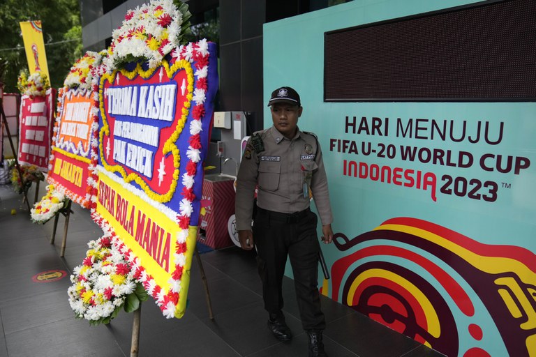 Seorang petugas berdiri di antara papan informasi pertandingan FIFA U-20 dan sejumlah karangan bunga tanda kekecewaan yang dikirim oleh para pendukung sepak bola di luar gedung PSSI di Jakarta, 30 Maret 2023. Sehari sebelumnya FIFA memituskan untuk membatalkan Indonesia sebagai tuan rumah Piala Dunia U-20, hanya delapan minggu sebelum dimulainya turnamen tersebut, menyusul gelombang penolakan sejumlah ormas Islam dan para politisi terkait partisipasi Israel dalam kejuaraan itu. [Dita Alangkara/AP Foto]