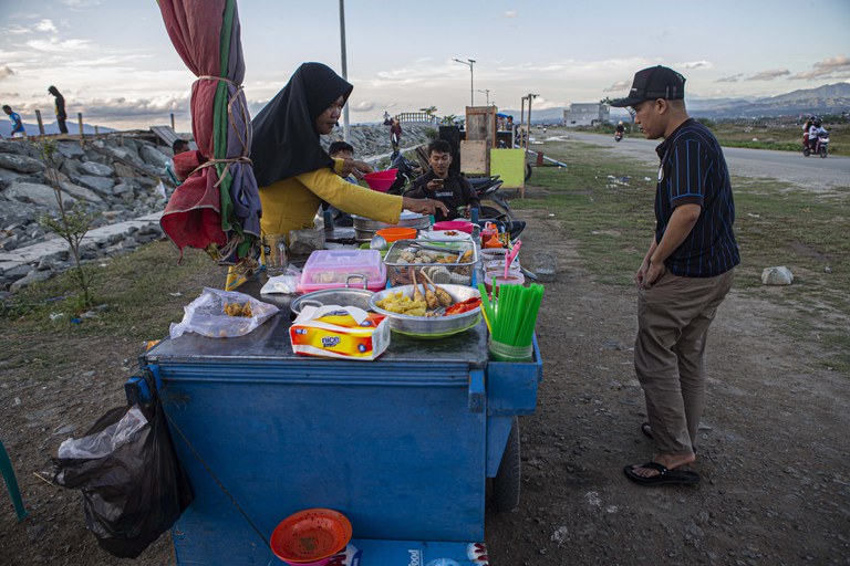 Seorang penyintas gempa bumi dan tsunami (kiri) melayani pembeli yang datang ke tempat usahanya di depan tanggul Teluk Palu di pantai penggaraman, Palu, Sulawesi Tengah, Sabtu, 18 Februari 2023. Selain berusaha di atas tanggul, banyak juga penyintas membuka usaha dengan berjualan aneka gorengan di depan tanggul. [Taufan Bustan/BenarNews]
